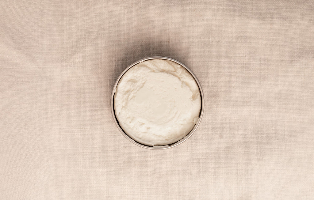 Open jar of tallow body creme on a beige background viewed from the top.
