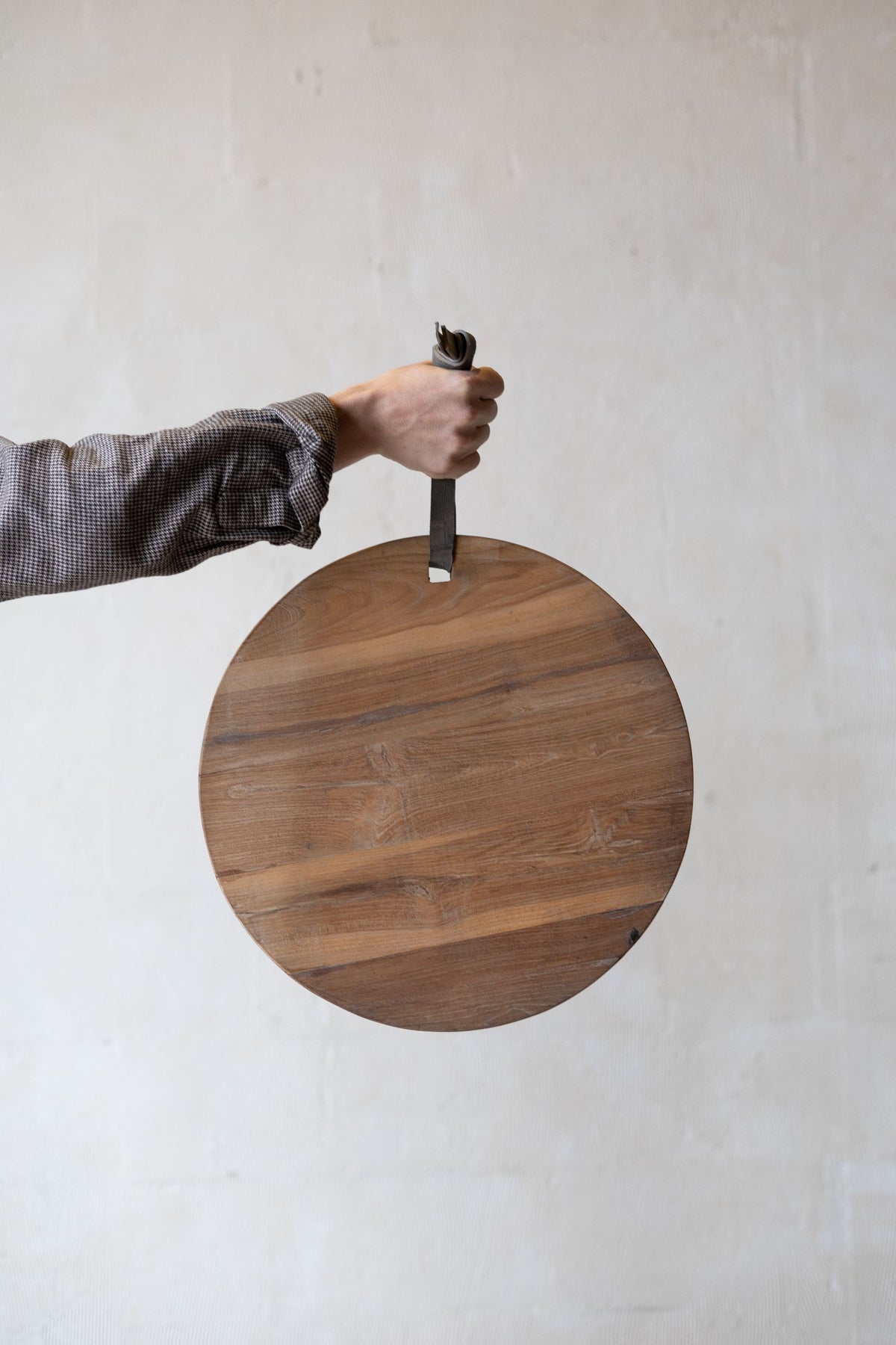 Round teak wood cheese board being held by leather strap. 