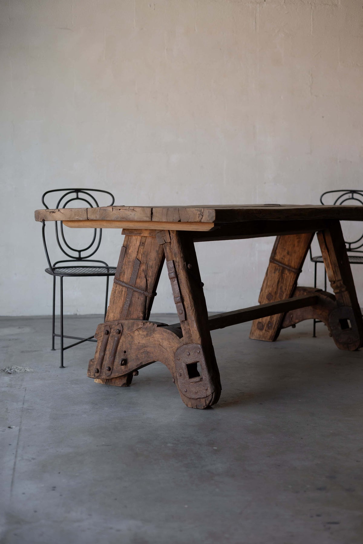 Rustic wooden sawhorse table with a thick, weathered tabletop and sturdy A-frame legs, featuring rustic metal reinforcements. Two art deco black iron chairs chairs with intricate backrest designs are placed beside the table, set against a minimalist concrete floor and plain wall background. Main image
