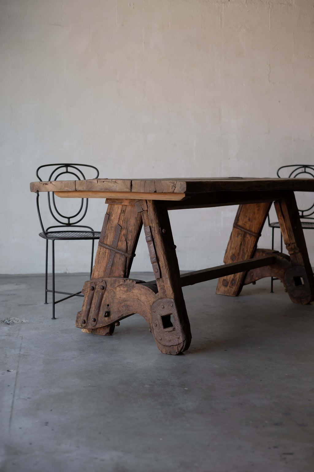 Rustic wooden sawhorse table with a thick, weathered tabletop and sturdy A-frame legs, featuring rustic metal reinforcements. Two art deco black iron chairs chairs with intricate backrest designs are placed beside the table, set against a minimalist concrete floor and plain wall background.