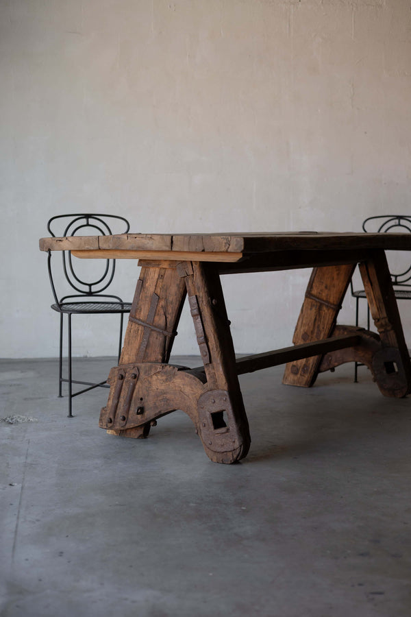 Rustic wooden sawhorse table with a thick, weathered tabletop and sturdy A-frame legs, featuring rustic metal reinforcements. Two art deco black iron chairs chairs with intricate backrest designs are placed beside the table, set against a minimalist concrete floor and plain wall background.