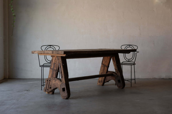 Rustic wooden dining table with a thick, weathered tabletop and sturdy A-frame legs made from reclaimed wood, featuring metal reinforcements. Two black metal chairs with intricate backrest designs are placed beside the table, set against a minimalist concrete floor and plain wall background.