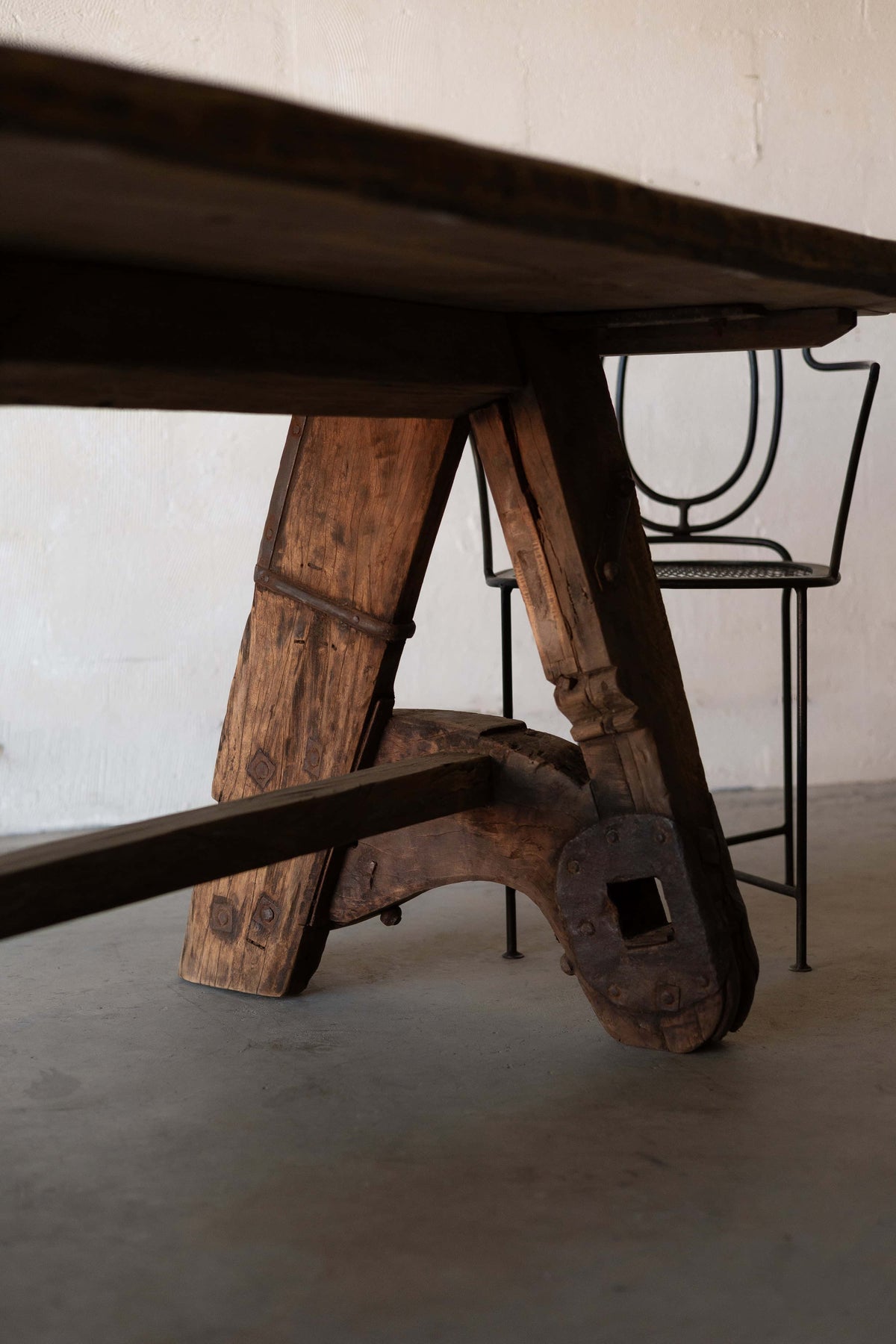 Close-up of rustic wooden dining table's A-frame, featuring industrial metal reinforcements. A black iron chair with art-deco backrest is placed behind the table.