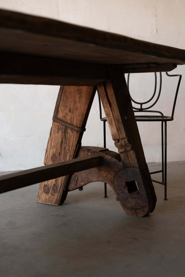 Close-up of rustic wooden dining table's A-frame, featuring industrial metal reinforcements. A black iron chair with art-deco backrest is placed behind the table.