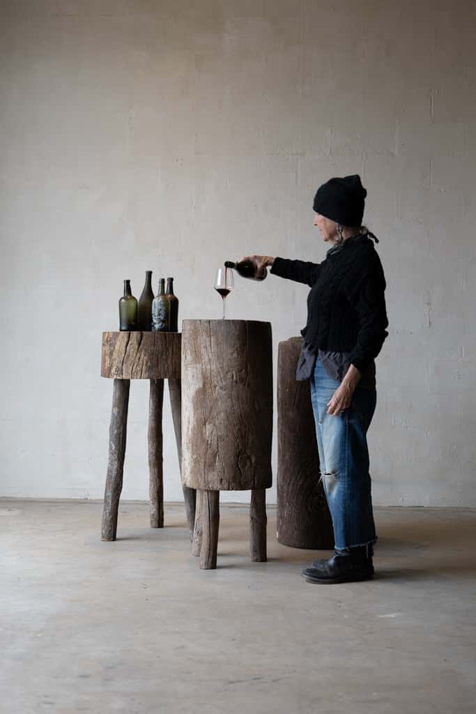 Woman pouring wine into a glass sitting on a butcher block table, with small table on the left holding four bottles of wine. 