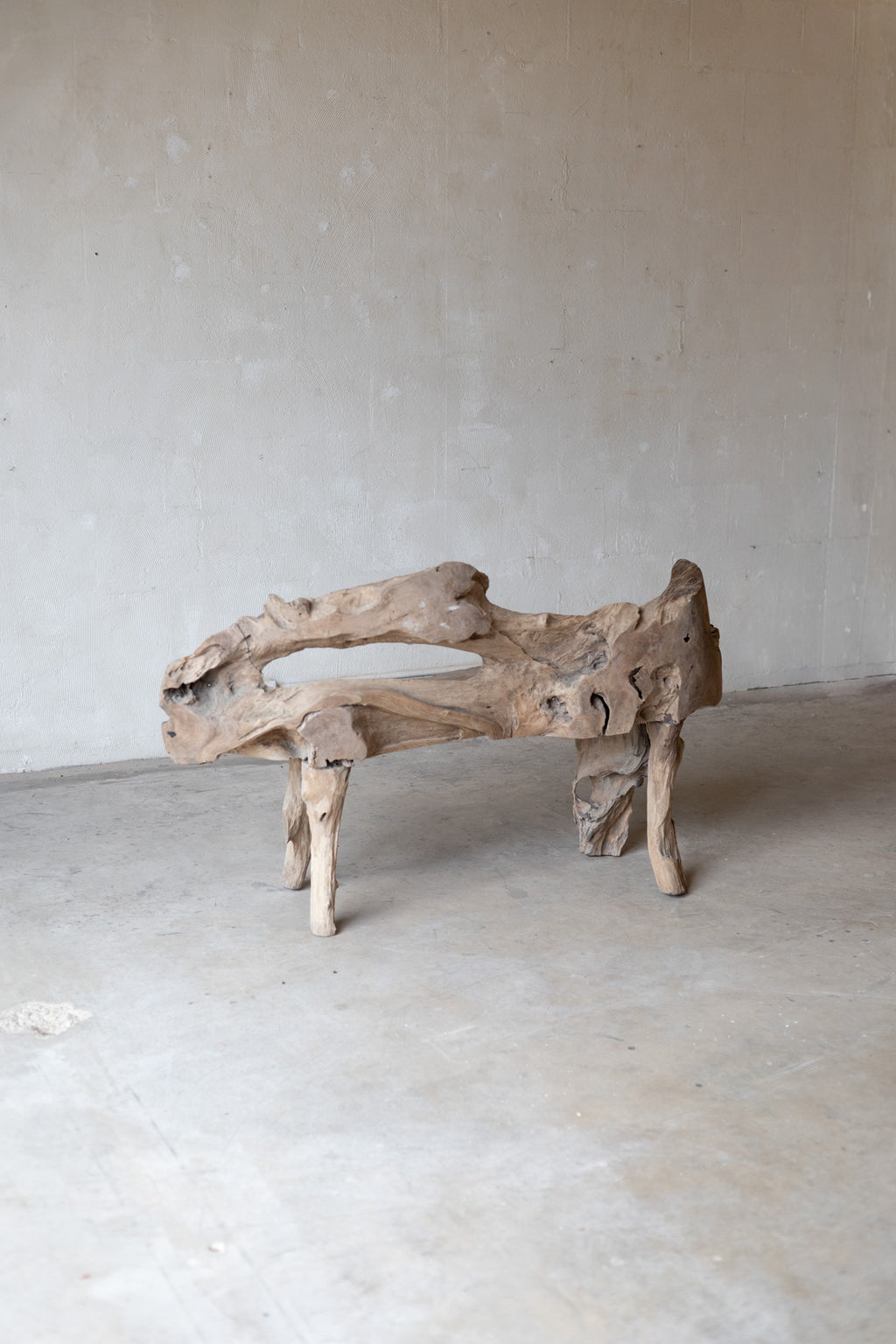 A rustic driftwood bench made from a natural log section, featuring a curved, hollow seat and four sturdy legs, set against a neutral concrete wall and floor.