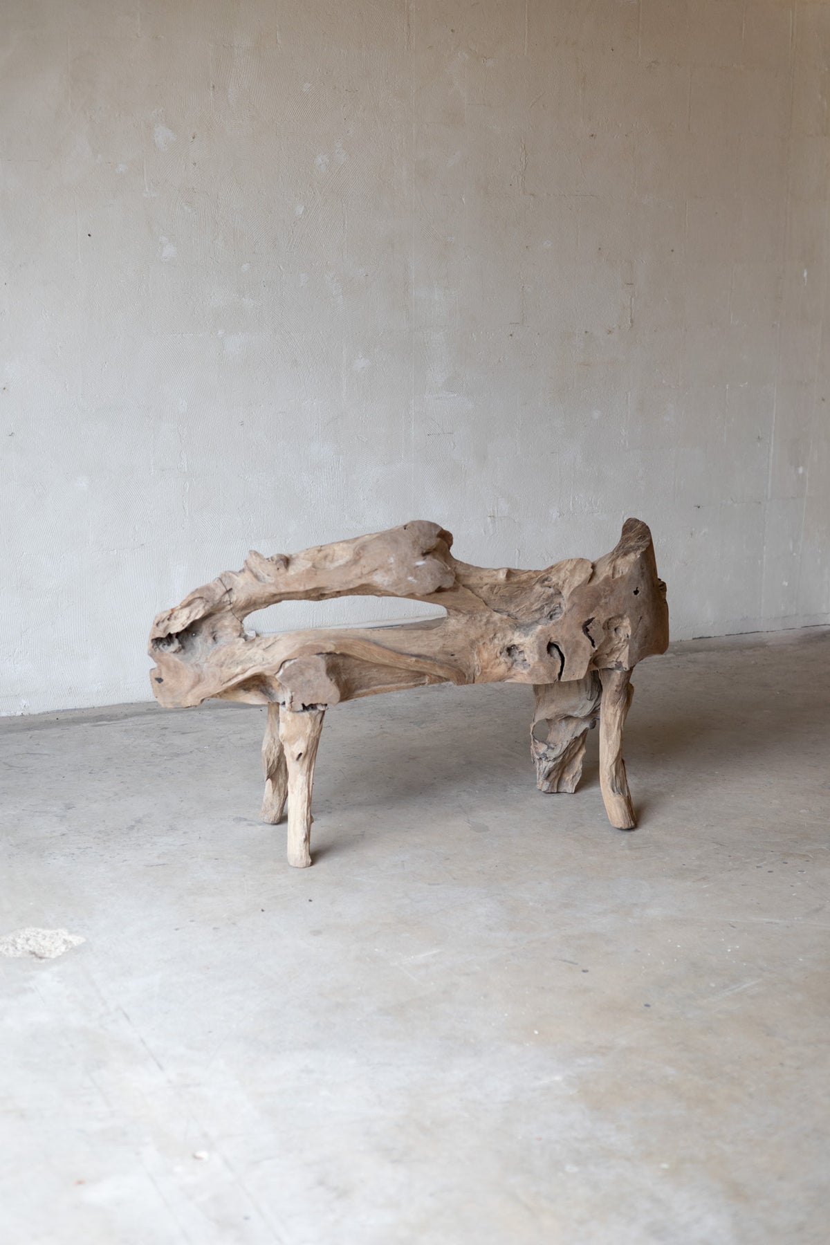 A rustic driftwood bench made from a natural log section, featuring a curved, hollow seat and four sturdy legs, set against a neutral concrete wall and floor.