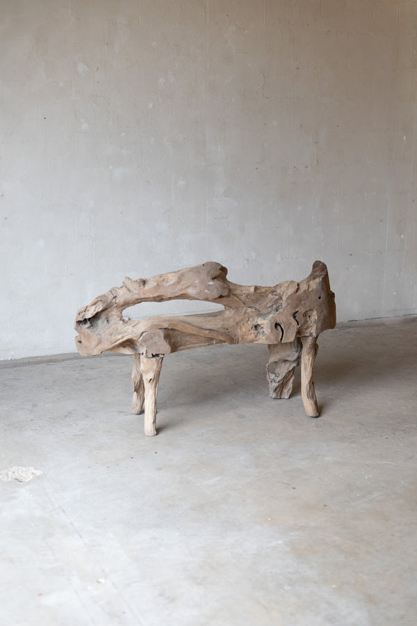 A rustic driftwood bench made from a natural log section, featuring a curved, hollow seat and four sturdy legs, set against a neutral concrete wall and floor.