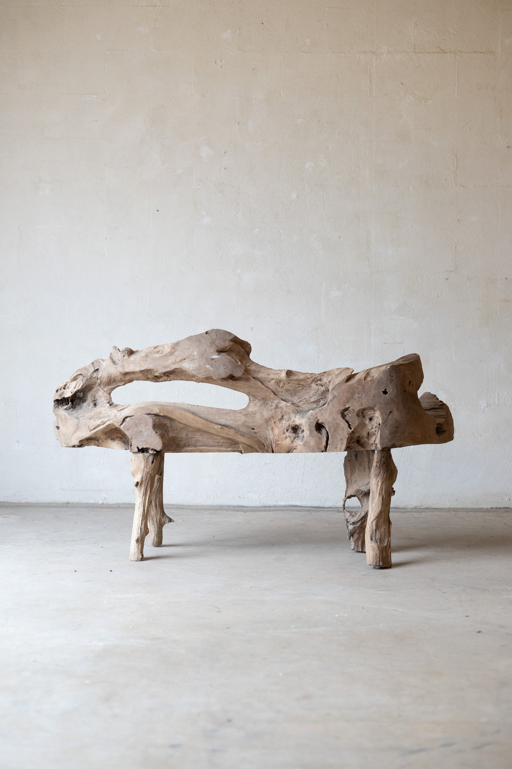 A rustic driftwood bench made from a natural irregular log with three short legs, photographed in a minimalist studio setting with a textured beige wall and concrete floor.