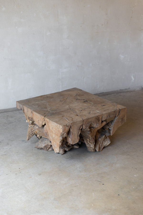 A rustic square wooden coffee table made from a natural tree stump with rough, uneven edges and visible grain, set against a plain concrete floor and wall.