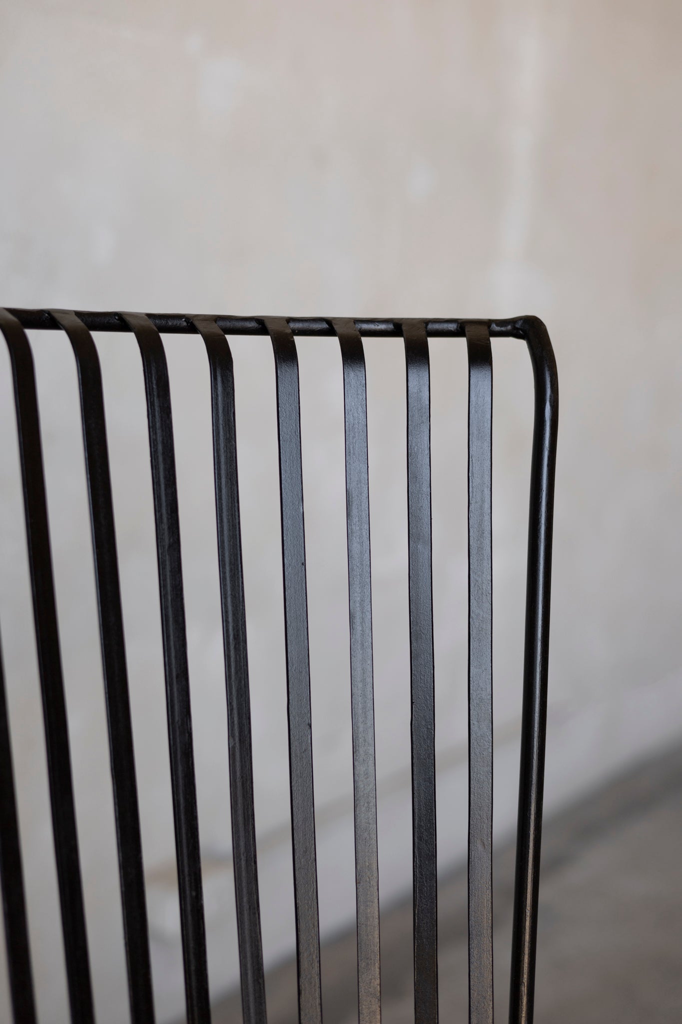 Close up of the corner of a black iron slatted chair against a blurred white background.