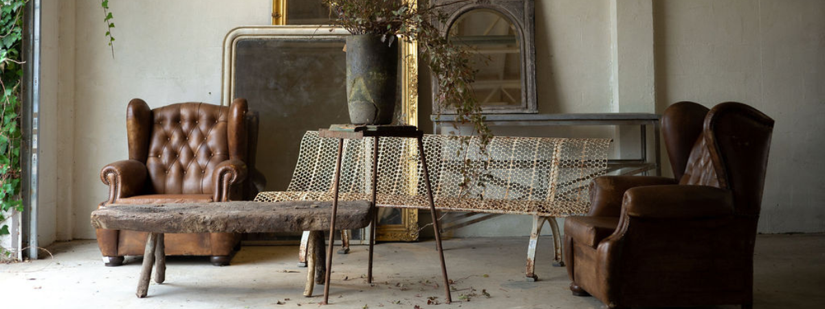 Vintage furniture and decor in a naturally lit room with beige walls featuring leather seats, mirrors and an iron bench. 