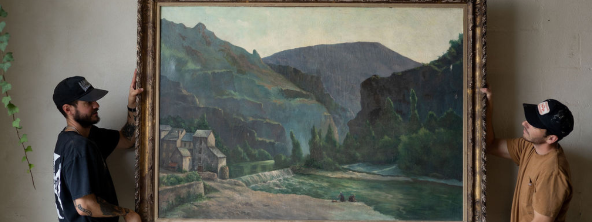 Two men hanging a large landscape painting on a beige wall.