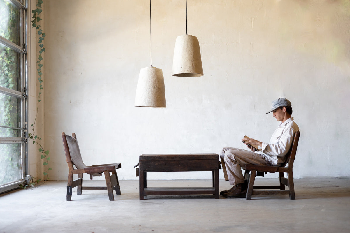 Person sitting on a wooden chair in a minimalistic room with large windows and textured white pendant lights.